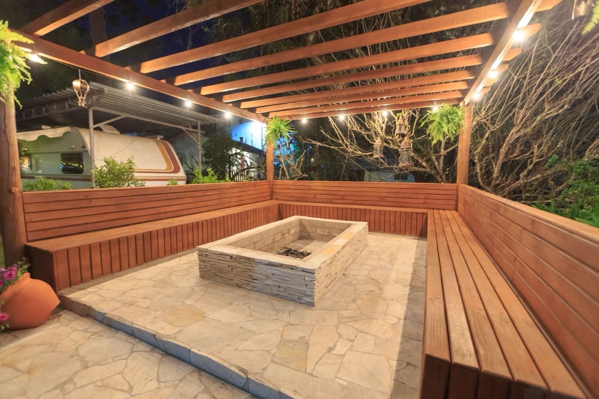 A lit pergola over a stone patio, with built-in bench seating around a fire pit.