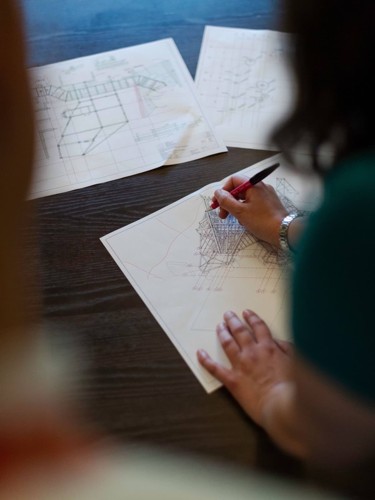 Hands with a red pen marking up structural drawings spread across a dark table.