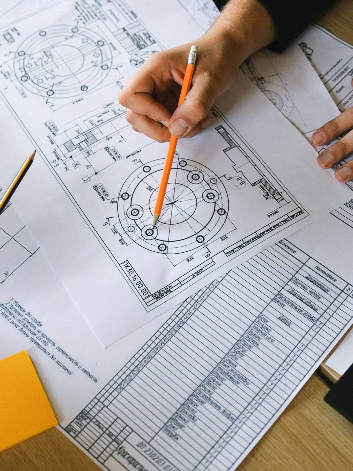 An orange pencil resting on an engineering drawing beside a printed parts table.