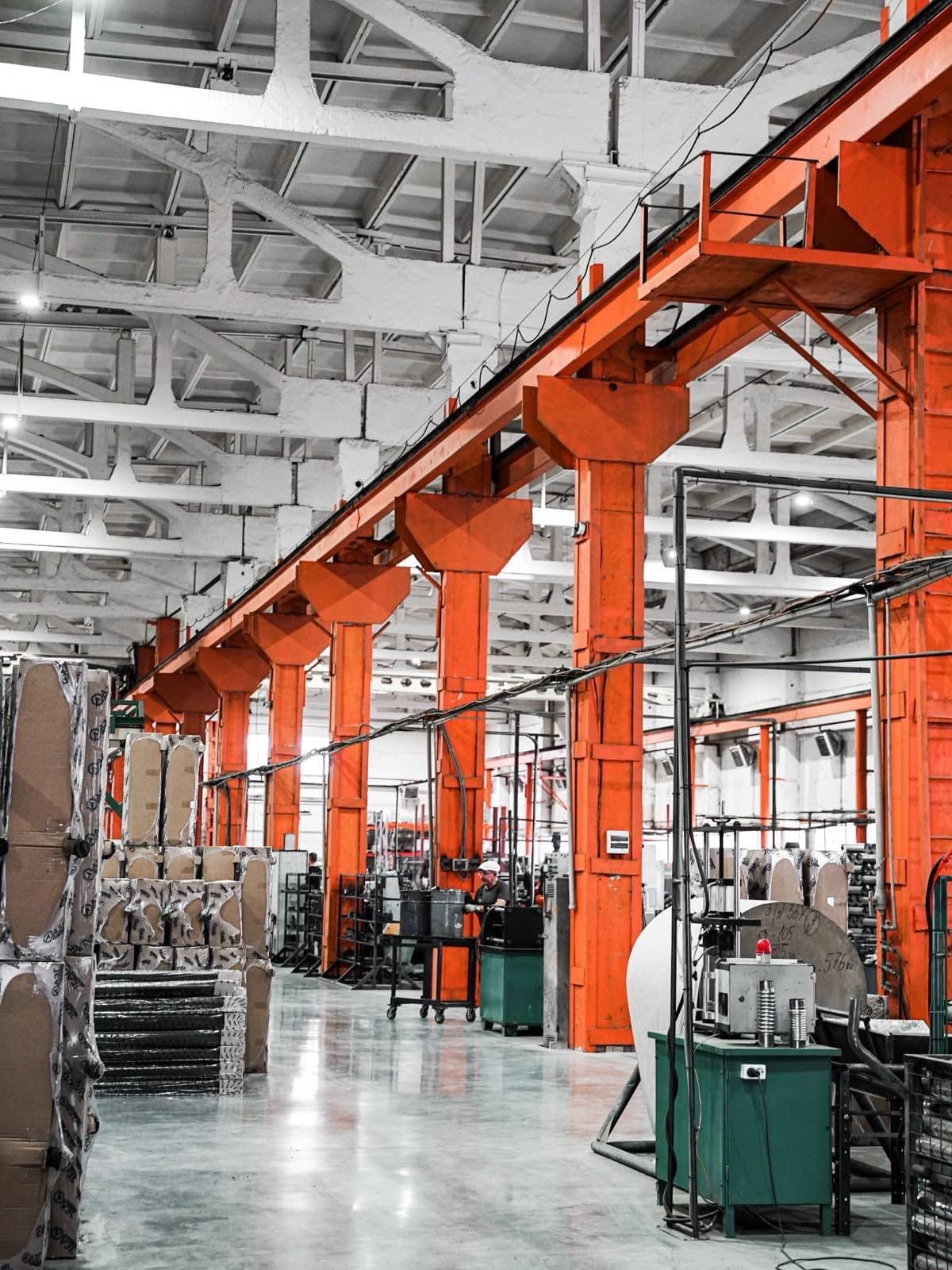 A factory floor under an orange steel gantry, one worker at a machine in the distance.