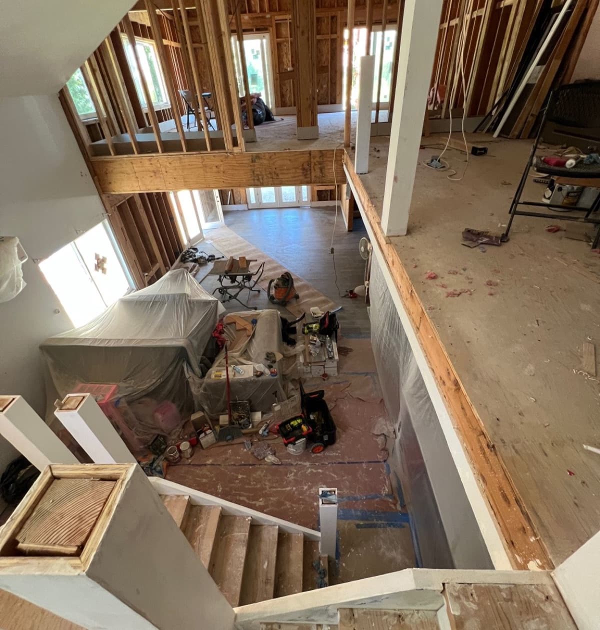 A gutted two-storey interior mid-remodel, seen from the stairs: bare framing, papered floors, and tools and materials staged across both levels.